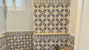 Et badeværelse med toilet, vask og bruseområde, udsmykket med mønstrede fliser og et vindue over bruseren.