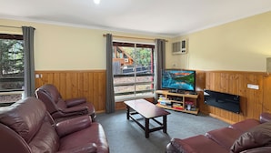 A living room with leather sofas, a wooden coffee table, a television on a wooden stand, and a window with a view of outdoor wooden structures. - Rosella Views - Cosy Cottage at the Base of Mt Buller (Sawmill Settlement)