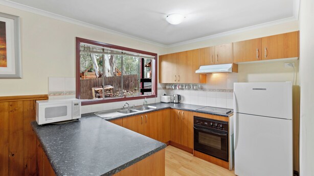 A kitchen with wooden cabinets, a black countertop, a white refrigerator, a stove, and a window with a view of trees. - Rosella Views - Cosy Cottage at the Base of Mt Buller (Sawmill Settlement)