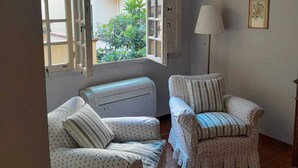 Una habitación con dos sillones tapizados, una lámpara de pie y un ventanal con contraventanas abiertas.