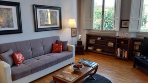Un salón con un sofá gris, almohadas rojas, una mesa de centro de madera con un cuenco, una estantería y una ventana con cortinas.