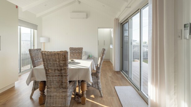 A dining area with wicker chairs, a white table, and a view of the outdoors through large windows.