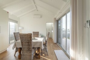 Un coin repas avec des chaises en osier, une table blanche et une vue sur l’extérieur grâce à de grandes fenêtres.