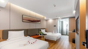 A hotel room with two beds, a small table with a flower arrangement, a painting on the wall, and a TV mounted on the wall.