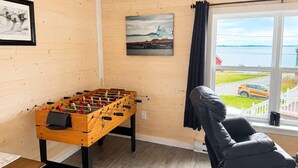 Una stanza con un tavolo da calcio in legno, una poltrona in pelle nera, una finestra che si affaccia su un lago e un'auto, e due quadri appesi al muro.