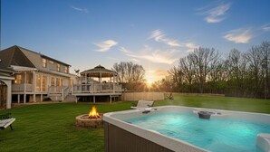 Un patio trasero con un jacuzzi, una fogata, una terraza cubierta y una casa grande.