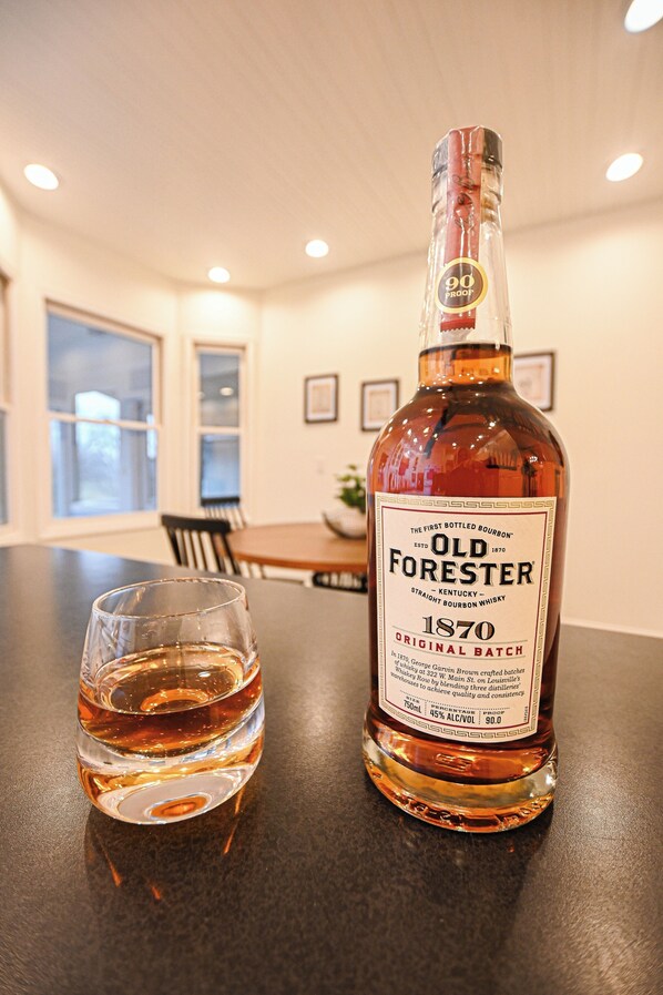 A bottle of Old Forester 1870 bourbon whiskey and a glass on a table in a room with a dining area in the background.