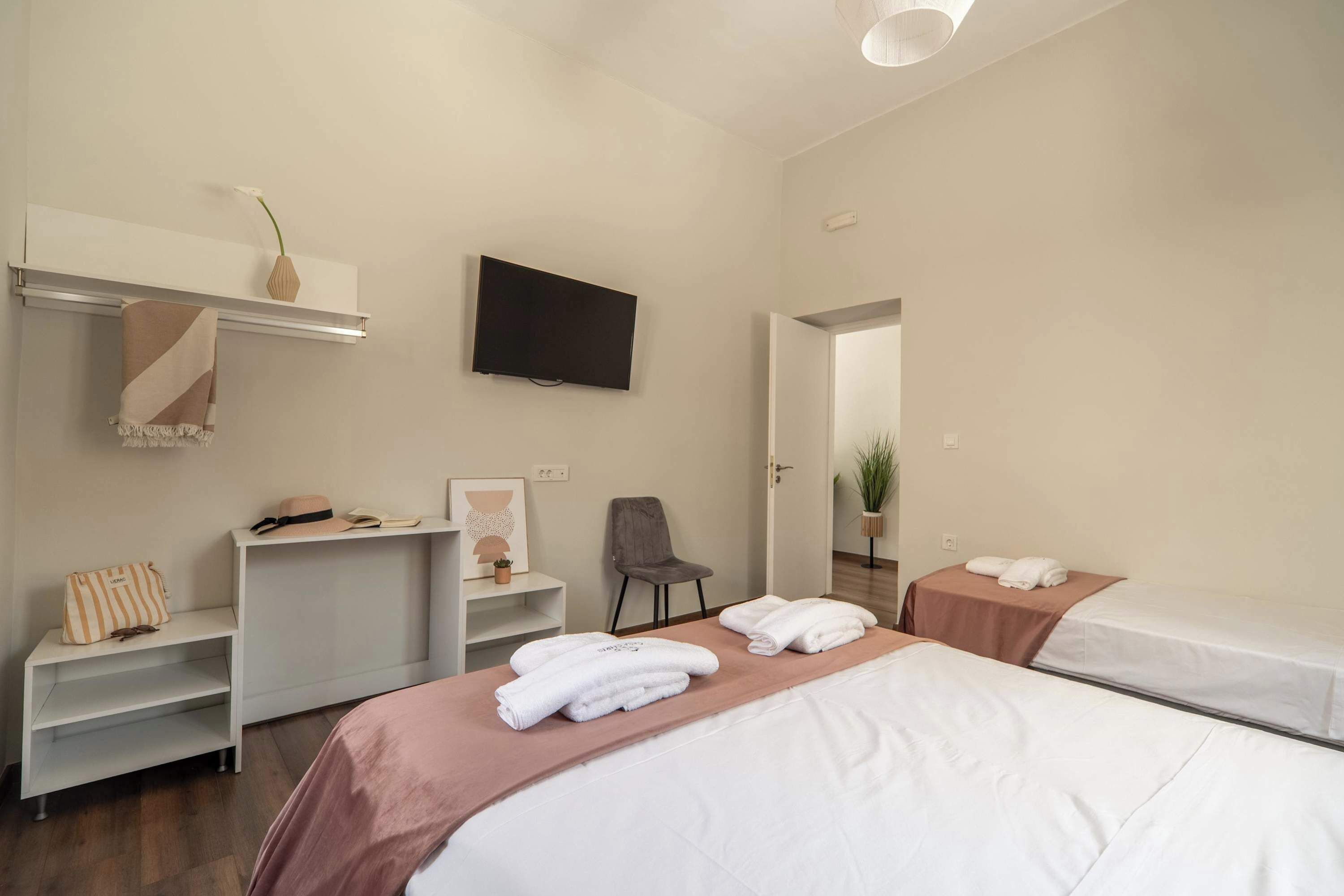 A hotel room with a bed, a TV, a chair, a desk, and a shelf with a vase and a hat.