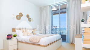 A modern bedroom with a large bed, a bedside table, and a view of the city. - Bloomfields 1BR The Pearl of Julphar (Abu Dabi)