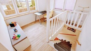 A room with a desk, a chair, a small table with toys, a staircase, and a window with blinds.