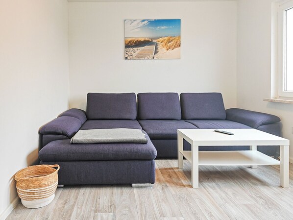 Una sala de estar con un sofá modular, una mesa de centro blanca, una cesta tejida y un cuadro con una escena de playa.