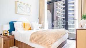 Een moderne slaapkamer met een groot bed, een nachtkastje, een bijzetmeubel, een raam met uitzicht op wolkenkrabbers en een kunstwerk in een lijst aan de muur.