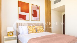 Quarto com cama, mesa de cabeceira branca e luminária. Há duas pinturas abstratas na parede.