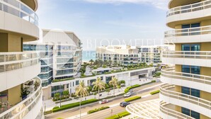 Un edificio moderno de varios pisos con balcones, una zona ajardinada con palmeras y una carretera en primer plano.