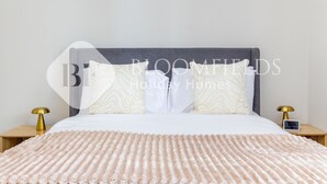 A neatly made bed with a patterned pillow, a bedside table with a lamp, and a wall-mounted clock.