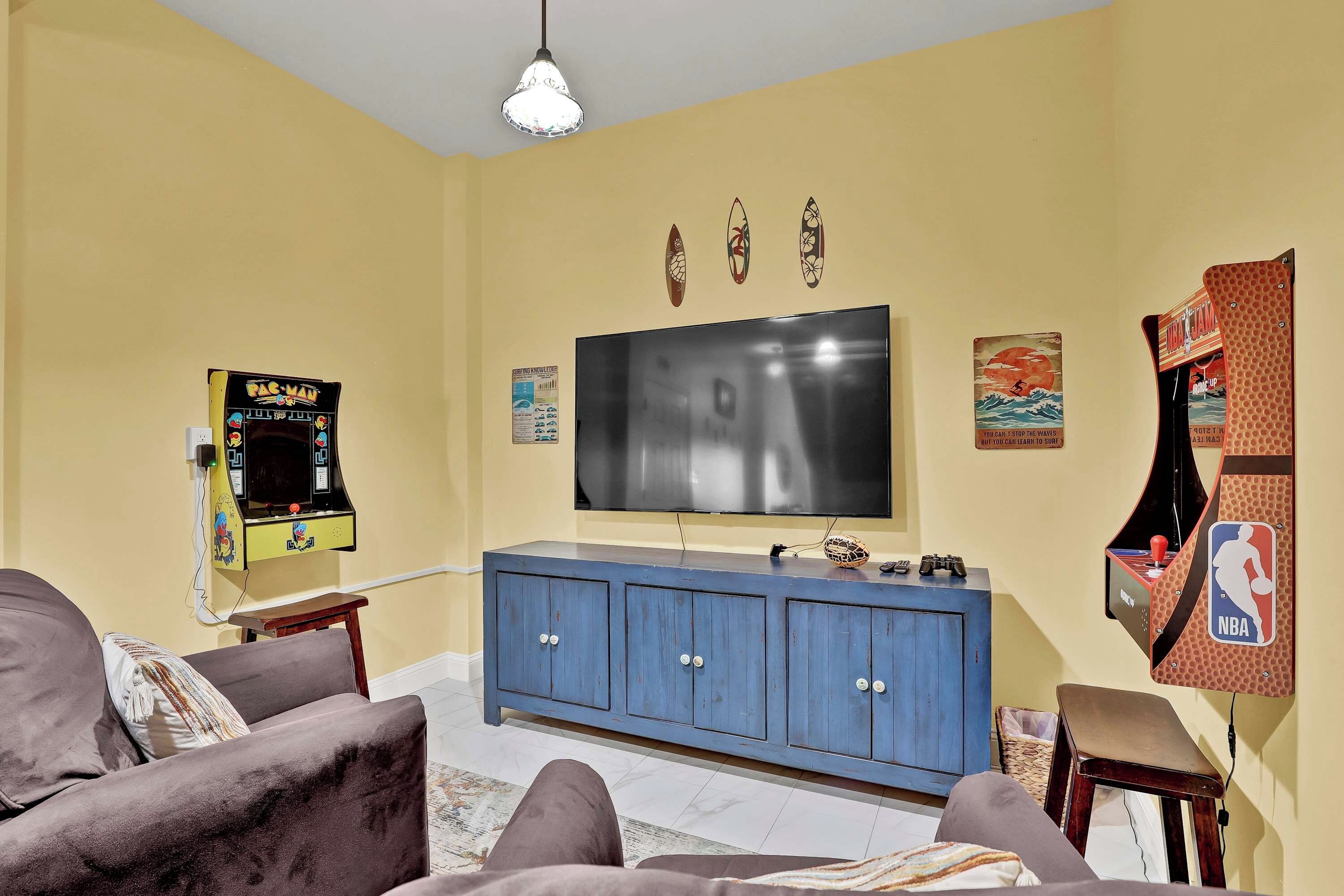 A living room with a flat-screen TV, a blue cabinet, a Pac-Man arcade machine, and a small table.