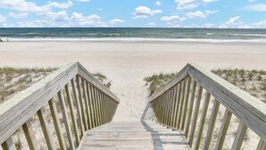 Ein hölzerner Steg führt zu einem Strand mit klarem Himmel und Blick auf das Meer.