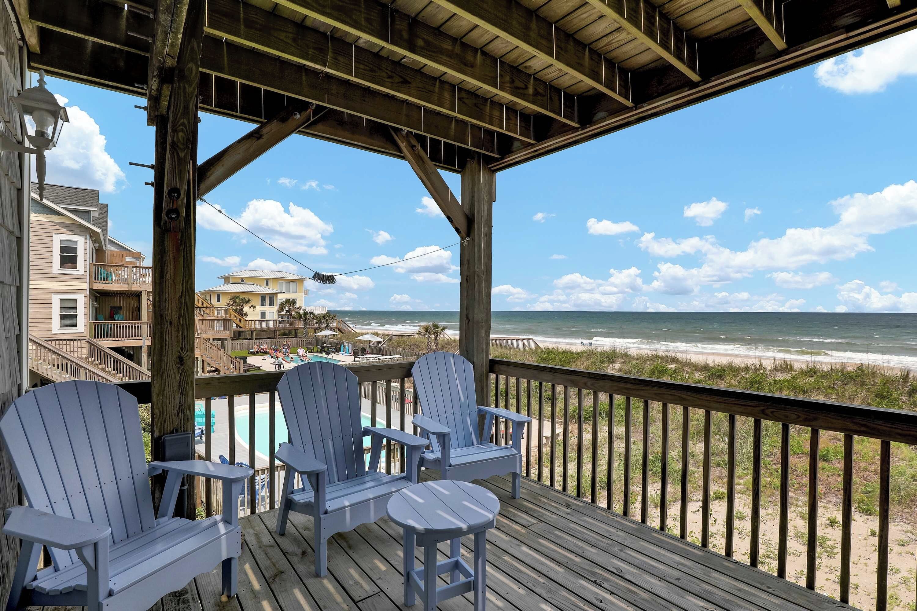 A wooden deck with two Adirondack chairs and a small table, overlooking a beach with a clear sky.
