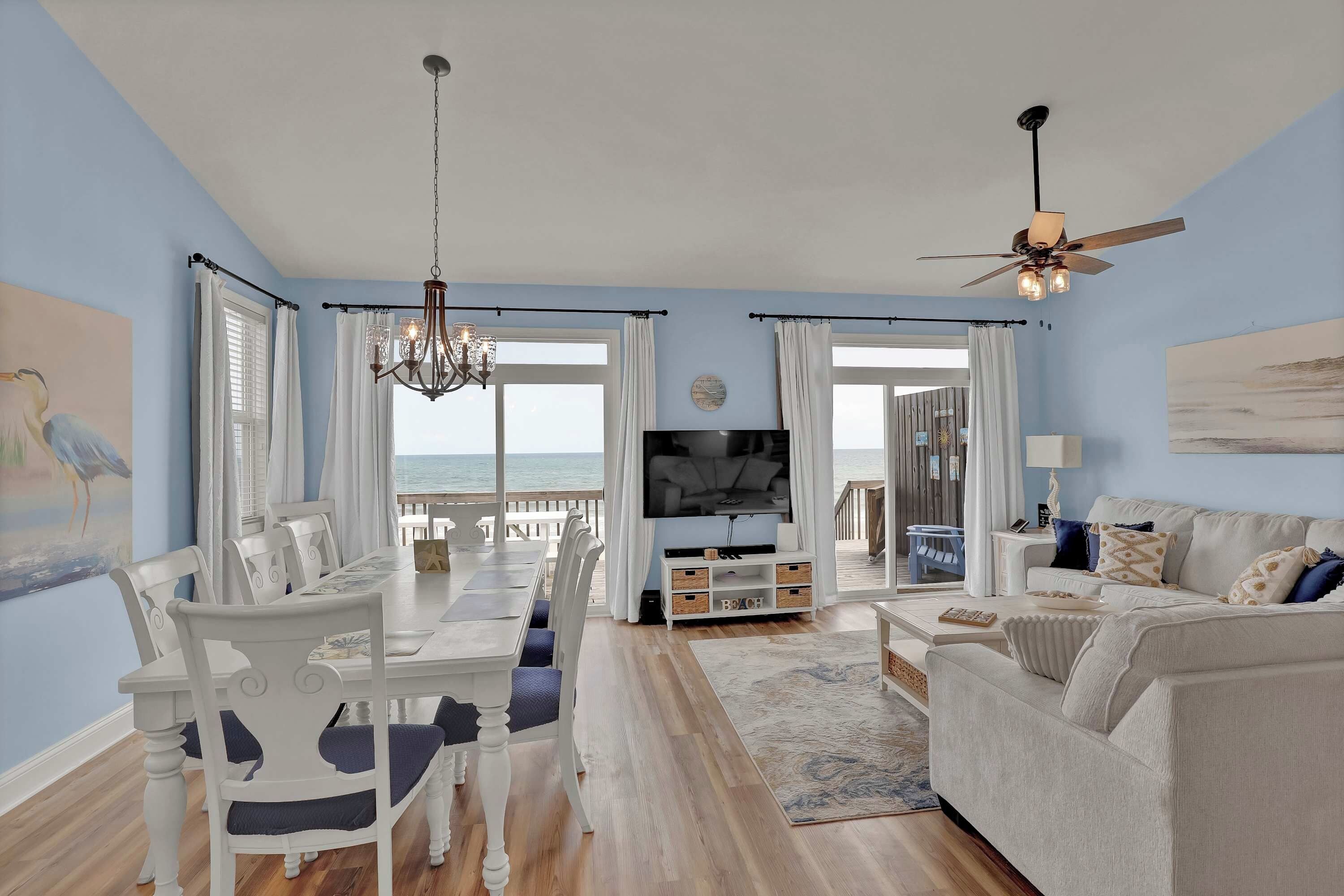 A living room with a TV, white furniture, a chandelier, and a view of the ocean.