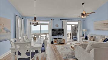 A living room with a TV, white furniture, a chandelier, and a view of the ocean.