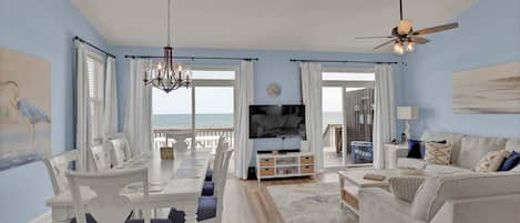 A living room with a TV, white furniture, a chandelier, and a view of the ocean.
