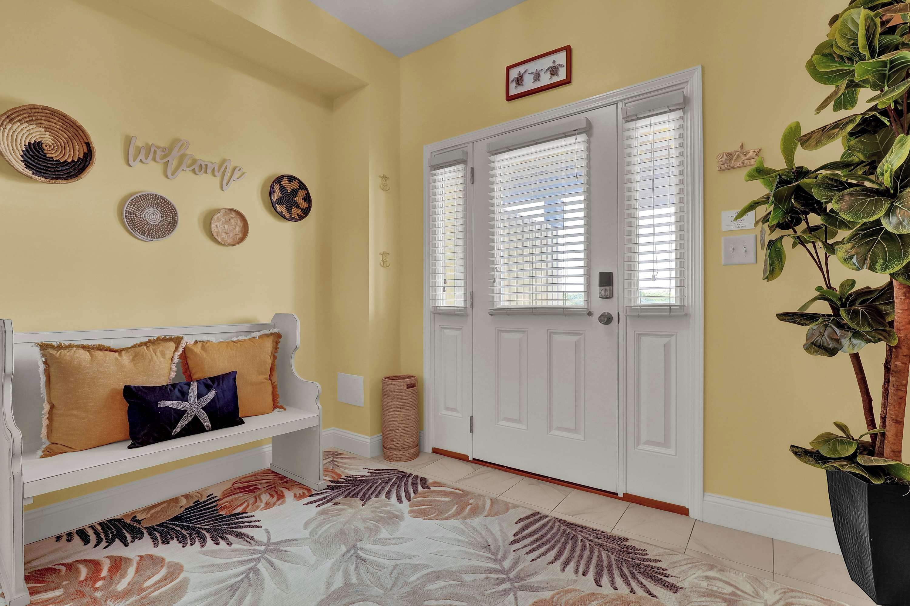 A hallway with a white bench, a welcome sign, decorative plates, and a door with blinds.