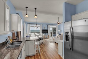 A modern kitchen with a stainless steel refrigerator, white cabinetry, and a wooden floor. There is a dining area with a table and chairs, and a view of the ocean through large windows.