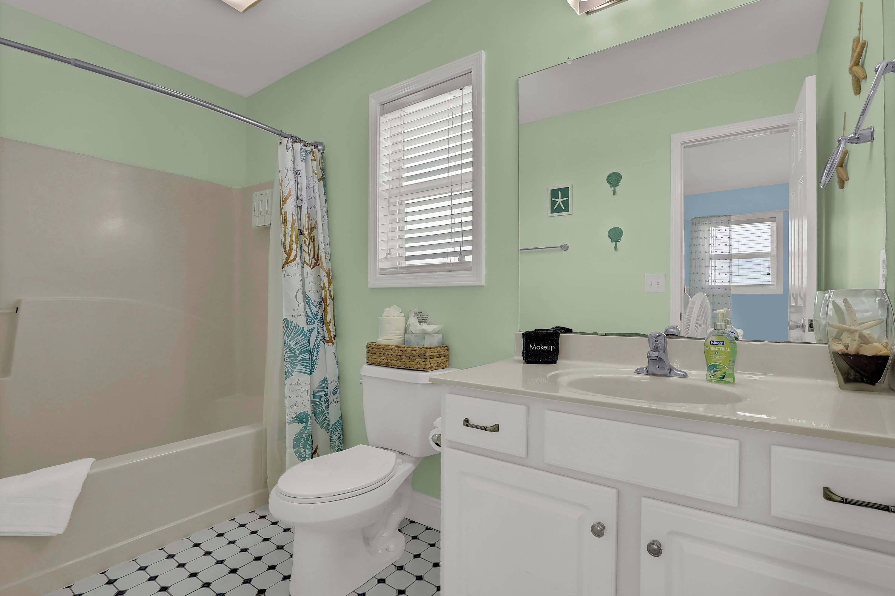 Une salle de bain avec des toilettes blanches, une baignoire, un rideau de douche orné de motifs de feuilles, un meuble-lavabo avec un miroir et un lavabo avec des articles de toilette.