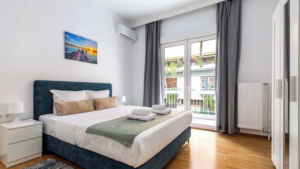 A modern bedroom with a large bed, a nightstand, a bedside table with a lamp, a painting on the wall, and a view of the outside balcony. - Cozyopolis - Timeless 3-BR in Athens  (Athens)