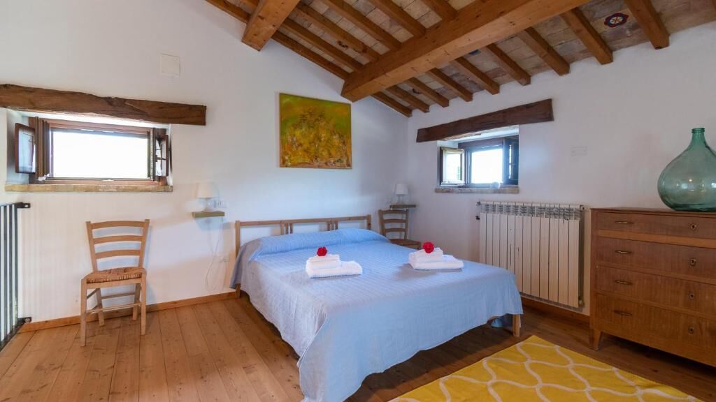 A bedroom with a wooden ceiling, a bed with a blue bedspread, two towels, a wooden chair, a dresser, and a radiator.
