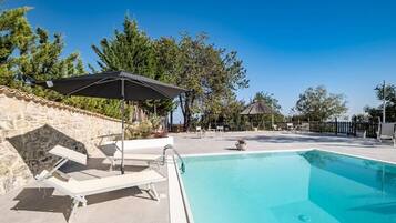 Ein Swimmingpool mit Liegestühlen und Sonnenschirm, umgeben von Steinmauern und Bäumen.