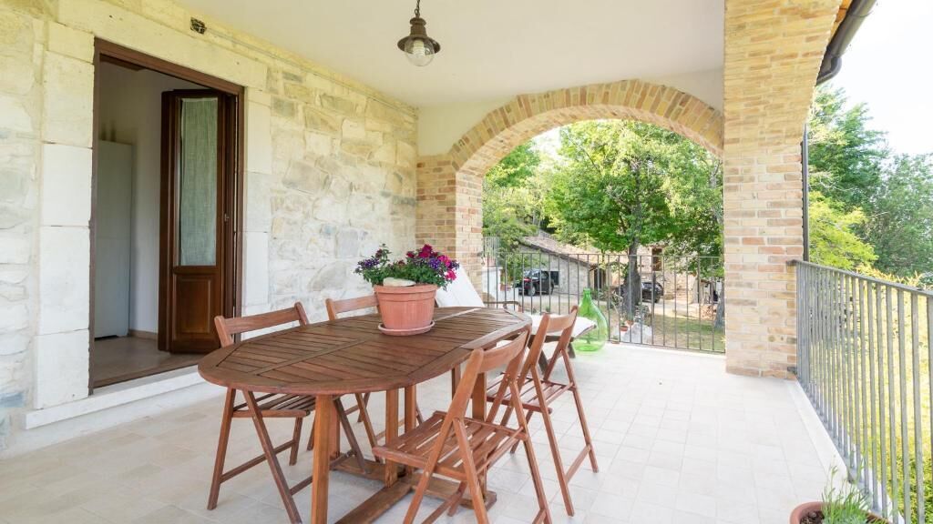 A wooden table with four chairs and a potted plant on a balcony with stone walls and a view of a tree and a fence.