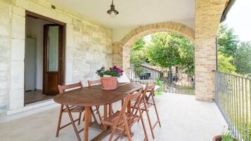 A wooden table with four chairs and a potted plant on a balcony with stone walls and a view of a tree and a fence.