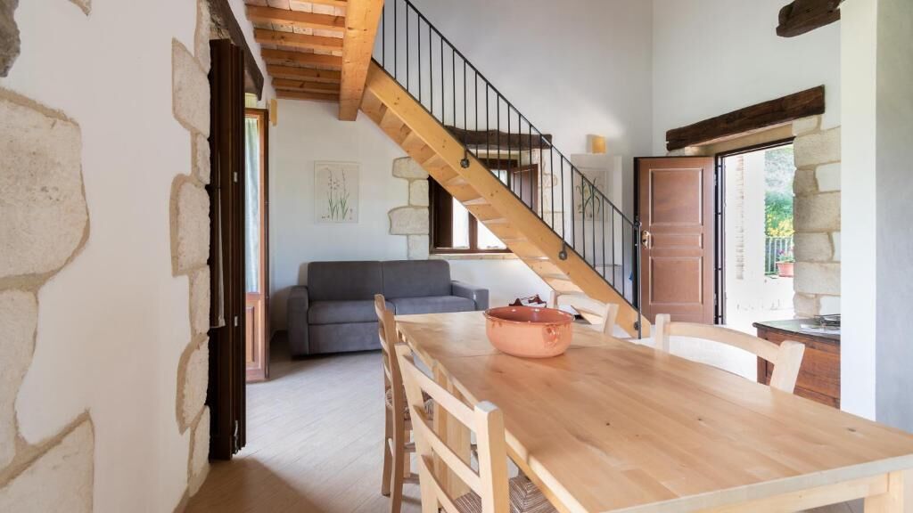 Un interno rustico con un tavolo da pranzo in legno, pareti in pietra, una scala e un divano grigio.