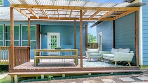 Une terrasse couverte avec un banc en bois et deux fauteuils en osier, une petite table et une porte donnant sur une maison aux murs recouverts de bardage bleu.