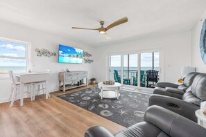 A living room with a dining area, a ceiling fan, a TV, and a view of the ocean. - Oceanfront condo w/ balcony & rocket launch views (Satellite Beach)
