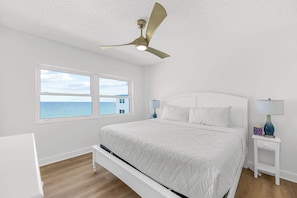 A bedroom with a bed, a ceiling fan, a nightstand with a lamp, and a window with a view of the ocean. - Oceanfront Condo w/ Balcony + Rocket Launch Views (Satellite Beach)
