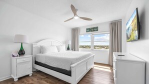 Une chambre à coucher comprenant un lit, une table de chevet, une lampe, un ventilateur de plafond, un téléviseur et une fenêtre donnant sur la plage.