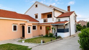 A two-story residential building with a red-tiled roof, a small front yard with grass and potted plants, and a visible chimney. - Apartment Olinda (Zaton)