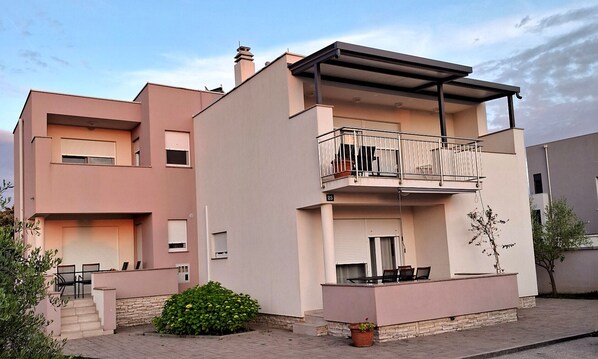 A two-story residential building with a balcony, a small garden, and a paved entrance.