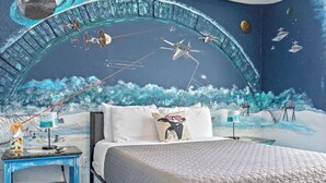 A bedroom with a mural of space-themed artwork, a bed with a quilted blanket, and a nightstand with a lamp.