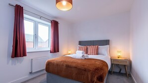 Ein ordentlich bezogenes Bett mit einer braunen Decke, einer Nachttischlampe und einem Fenster mit Vorhängen.