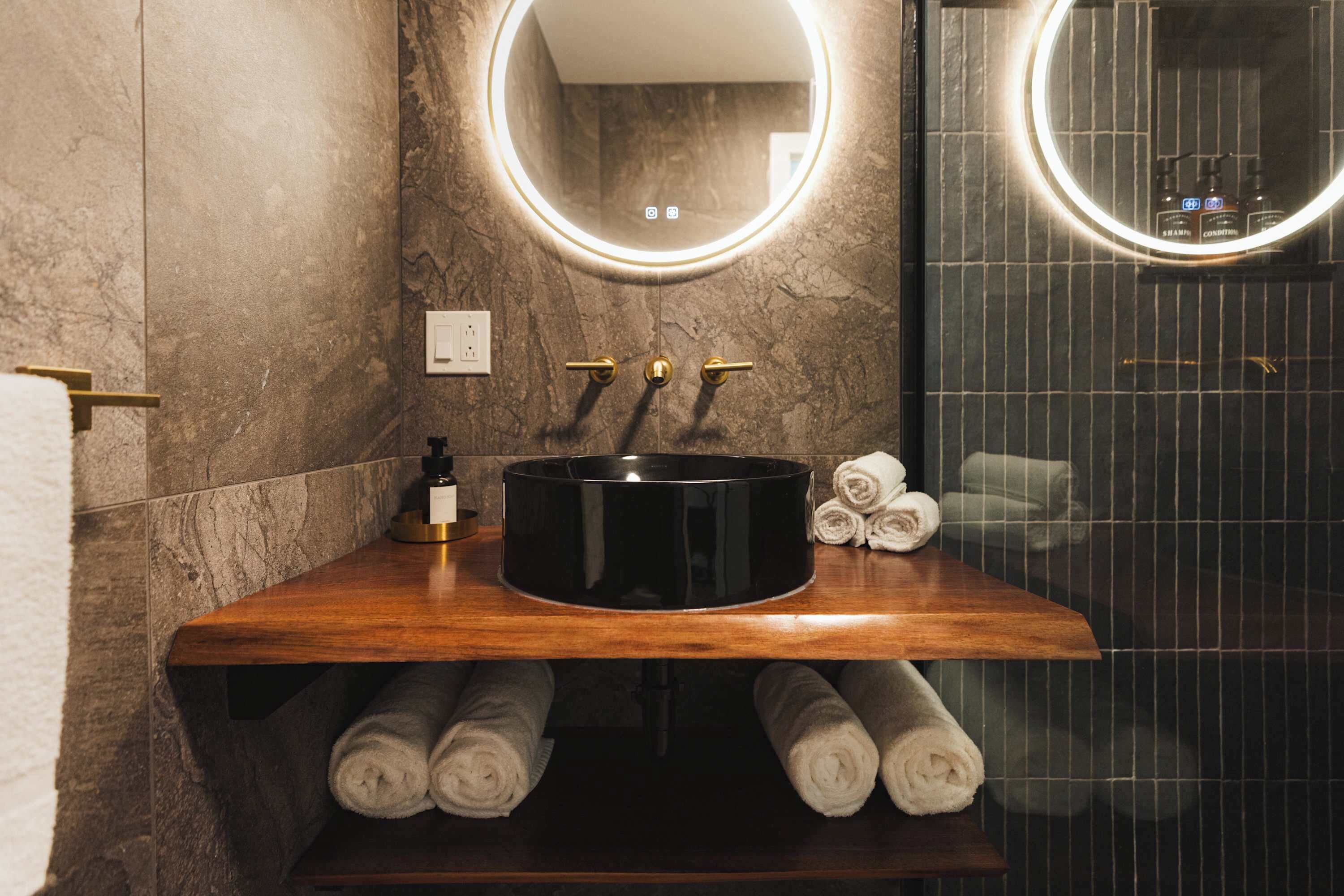 A bathroom with a wooden vanity, a black sink, and a mirror with a circular light.