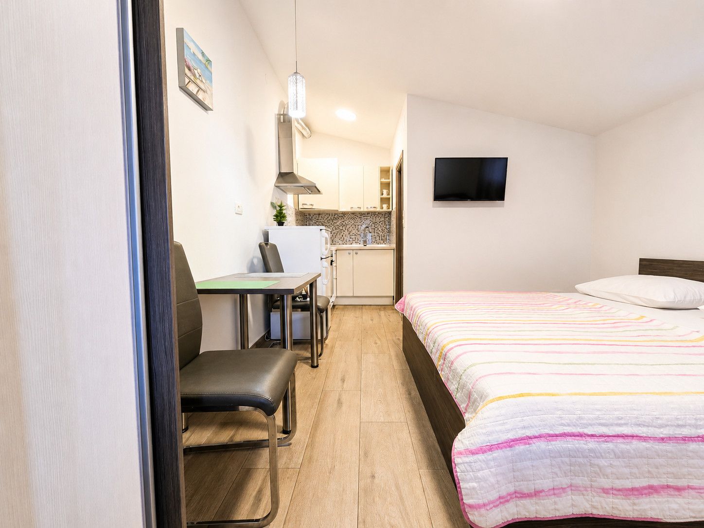 Une chambre d’hôtel compacte comprenant un lit, un bureau avec une chaise, une kitchenette et un téléviseur fixé au mur.