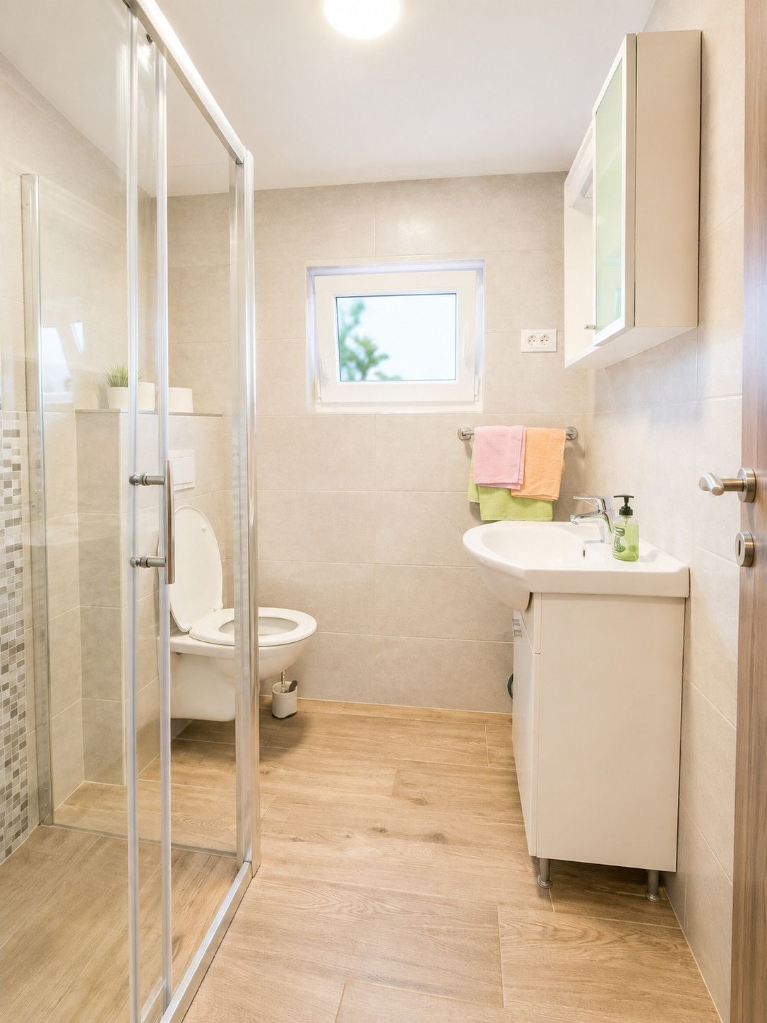 A modern bathroom with a glass shower enclosure, a toilet, a sink with a mirror above, and a small window providing natural light.