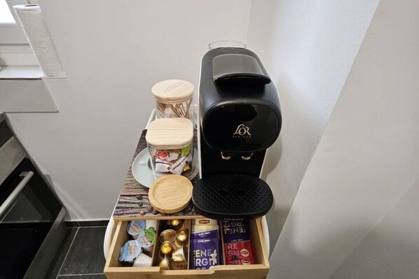 A compact kitchen area with a coffee maker, a wooden storage box with tea and snacks, and a small stool.