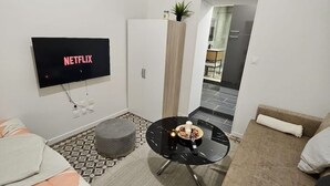A modern living room with a flat-screen TV displaying 'NETFLIX', a white wardrobe, a round black coffee table with a candle, and a patterned rug.