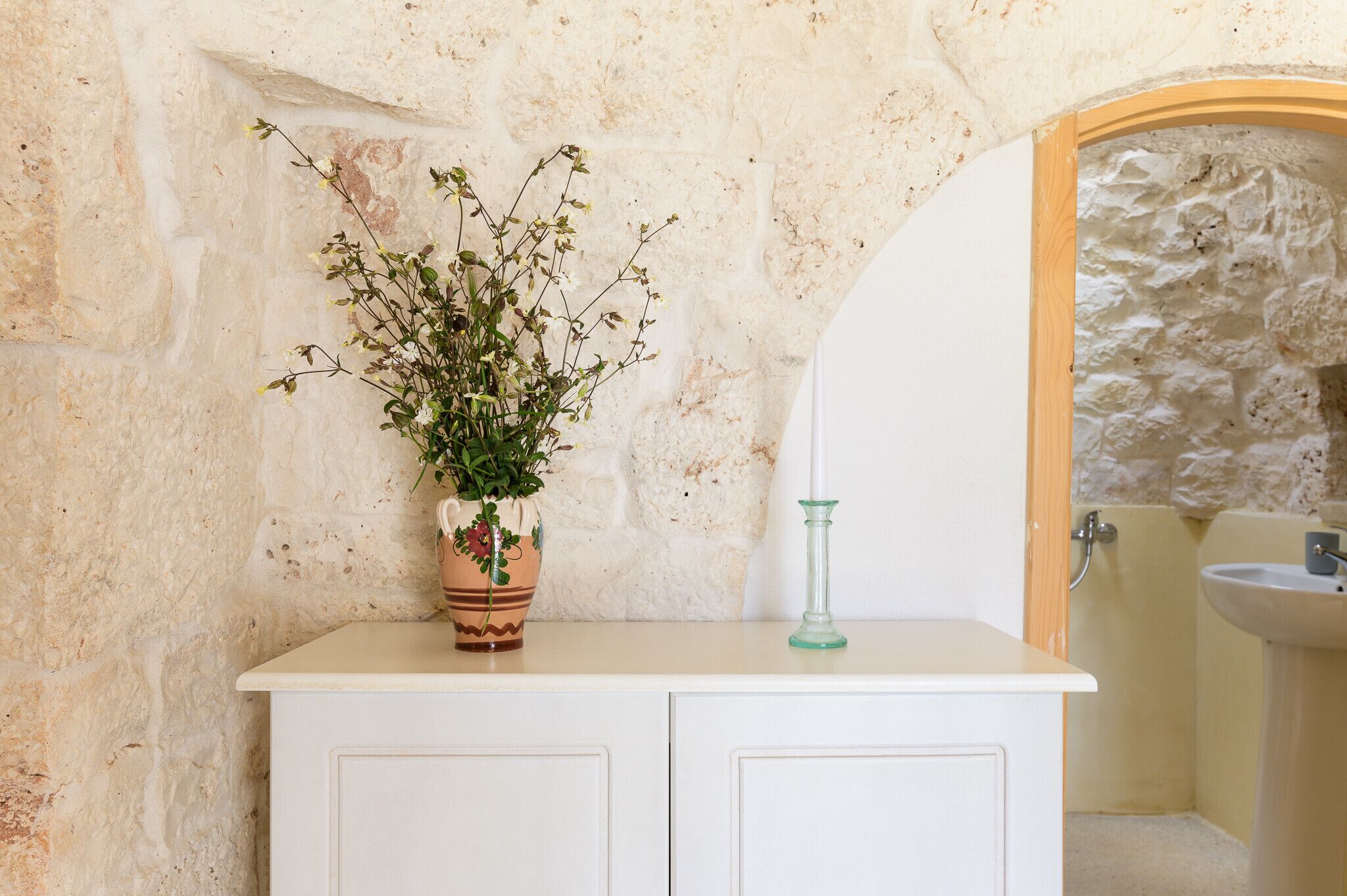 Une salle de bain avec un mur en pierre, un meuble blanc, une plante en pot et un porte-bougie en verre.