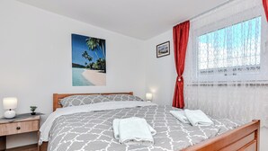 A bedroom with a wooden bed, a nightstand with a lamp, a painting of a beach scene, and a window with red curtains. - Apartments Ladika (Vir)