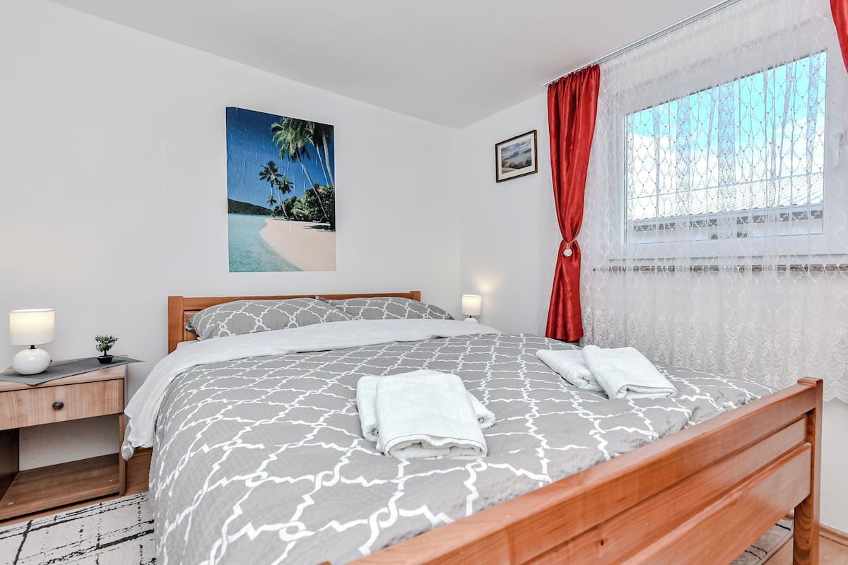 A bedroom with a wooden bed, a nightstand with a lamp, a painting of a beach scene, and a window with red curtains.